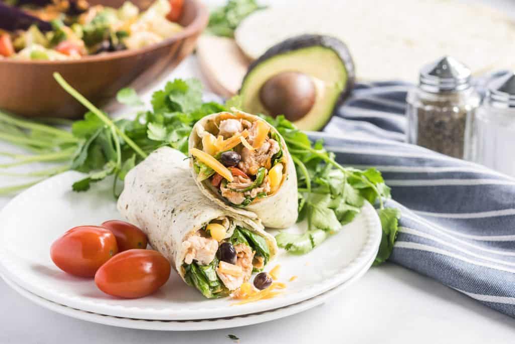 A BBQ Chicken Salad Wrap cut in half and stacked on a white plate with grape tomatoes and cilantro.