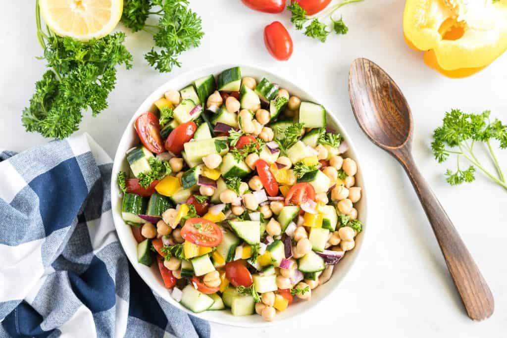 A white bowl of Chickpea Salad with a wooden spoon and ingredients scattered around it.