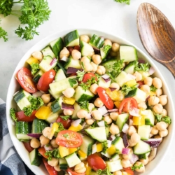 A white serving bowl full of Chickpea Salad with a wooden spoon and ingredients scattered around.