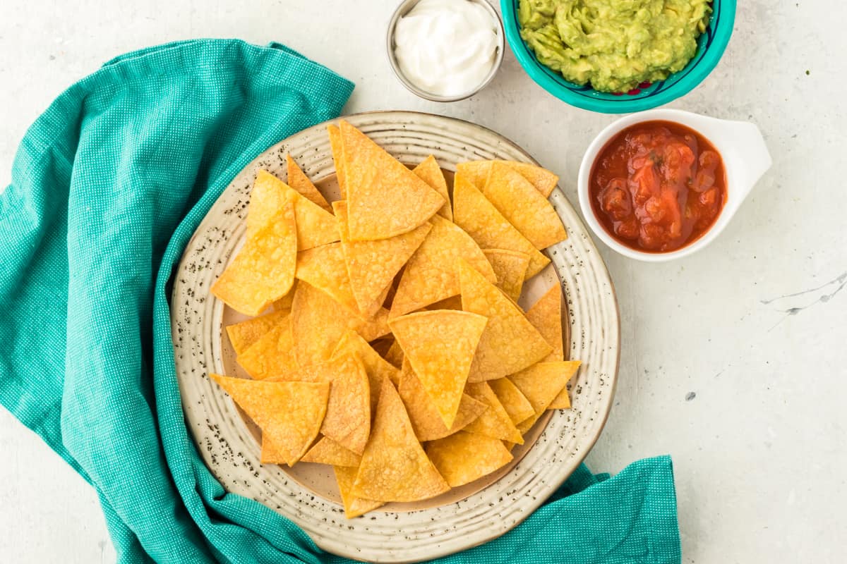 Air fryer tortilla chips on a plate with salsa , guacamole and sour cream on the side in bowls.