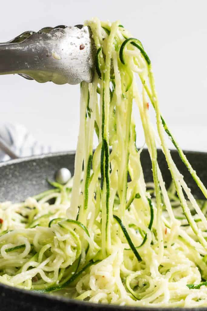 Metal tongs pull zucchini noodles out of a skillet.