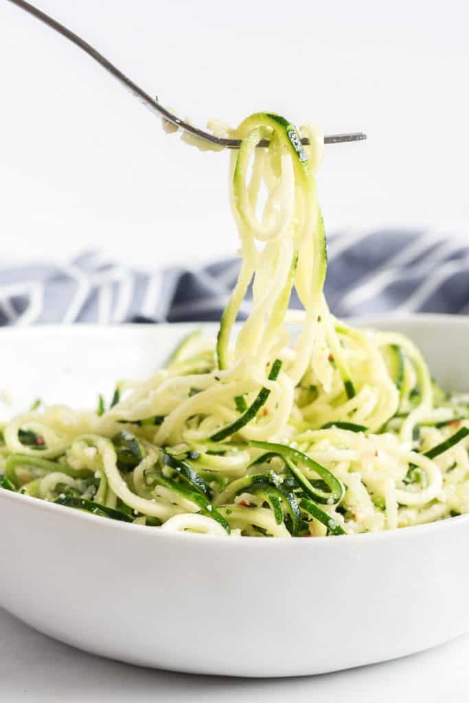 A fork lifts zucchini noodles from a white bowl.