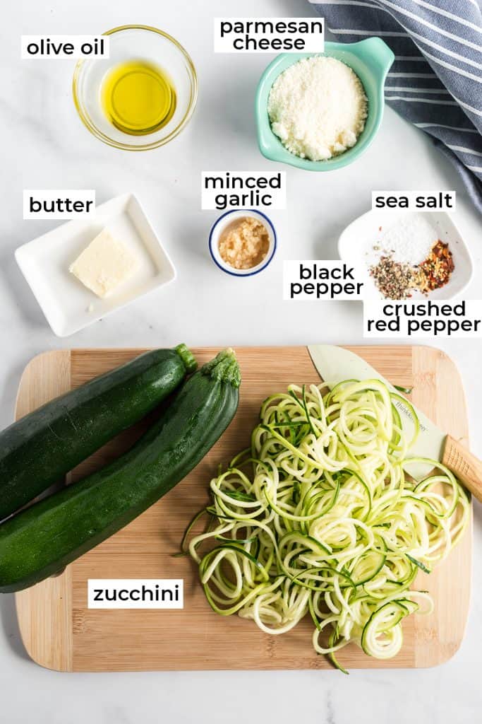 Zucchini, butter, olive oil and other ingredient for zoodles on a white surface.