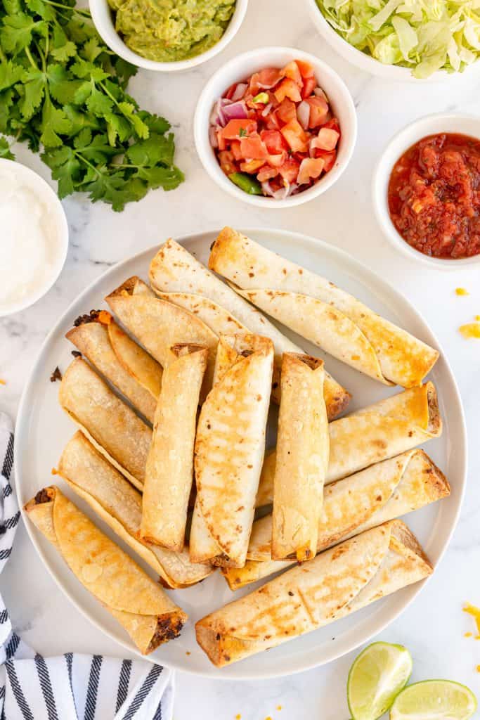 A platter of air fryer taquitos with toppings around it.