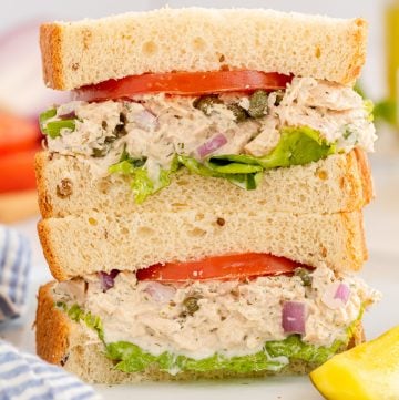 A tuna sandwich with tomatoes and lettuce cut in half and stacked on a plate.