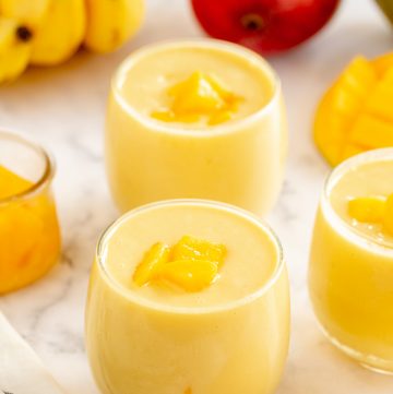 Two mango smoothies with honey, bananas, and a mango in the background.