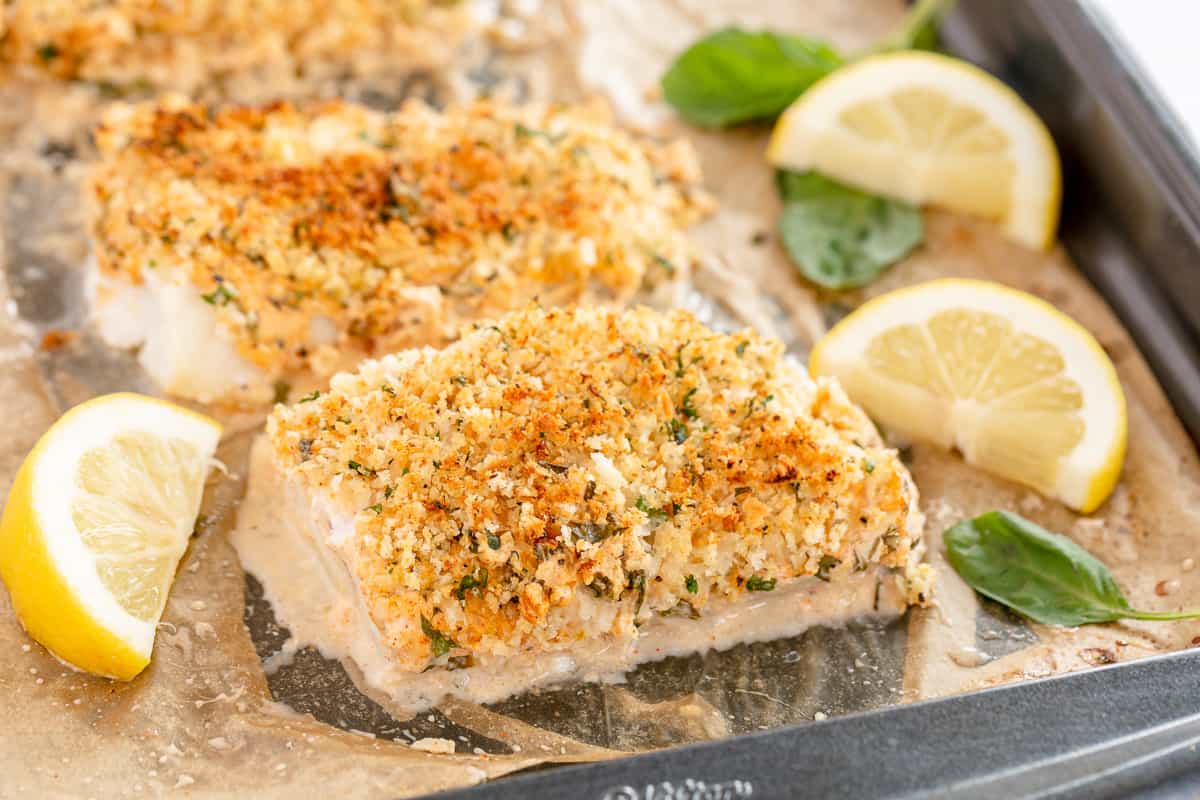 Close-up of a baking tray containing breaded and baked fish fillets garnished with fresh basil leaves and accompanied by lemon wedges. The fillets have a golden crust and sit on a parchment-lined tray.