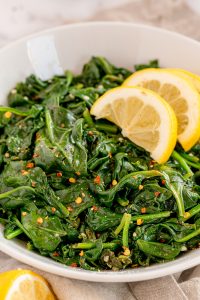 Sauteed spinach with crushed red pepper in a white bowl with lemon wedges.