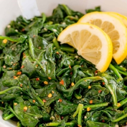 Sauteed spinach with crushed red pepper in a white bowl with lemon wedges.