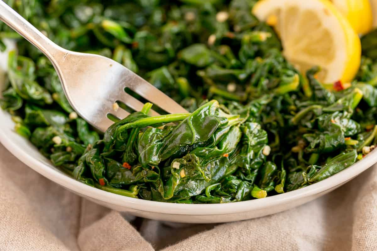 A close-up of a dish of sautéed spinach in a white bowl. A fork in the foreground holds a portion of spinach. Lemon slices are partially visible on the edge of the bowl. The spinach appears to be seasoned with visible spices.