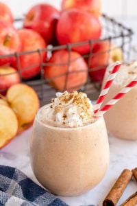 An apple pie smoothie with red and white straws in front of a crate of apples.