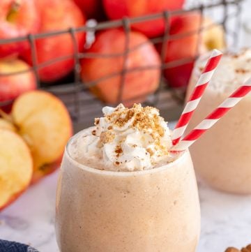 An apple pie smoothie with red and white straws in front of a crate of apples.
