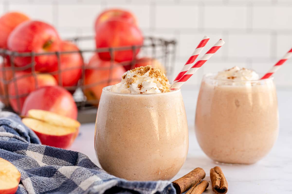 Two smoothies with red and white straws in front of a crate of apples.