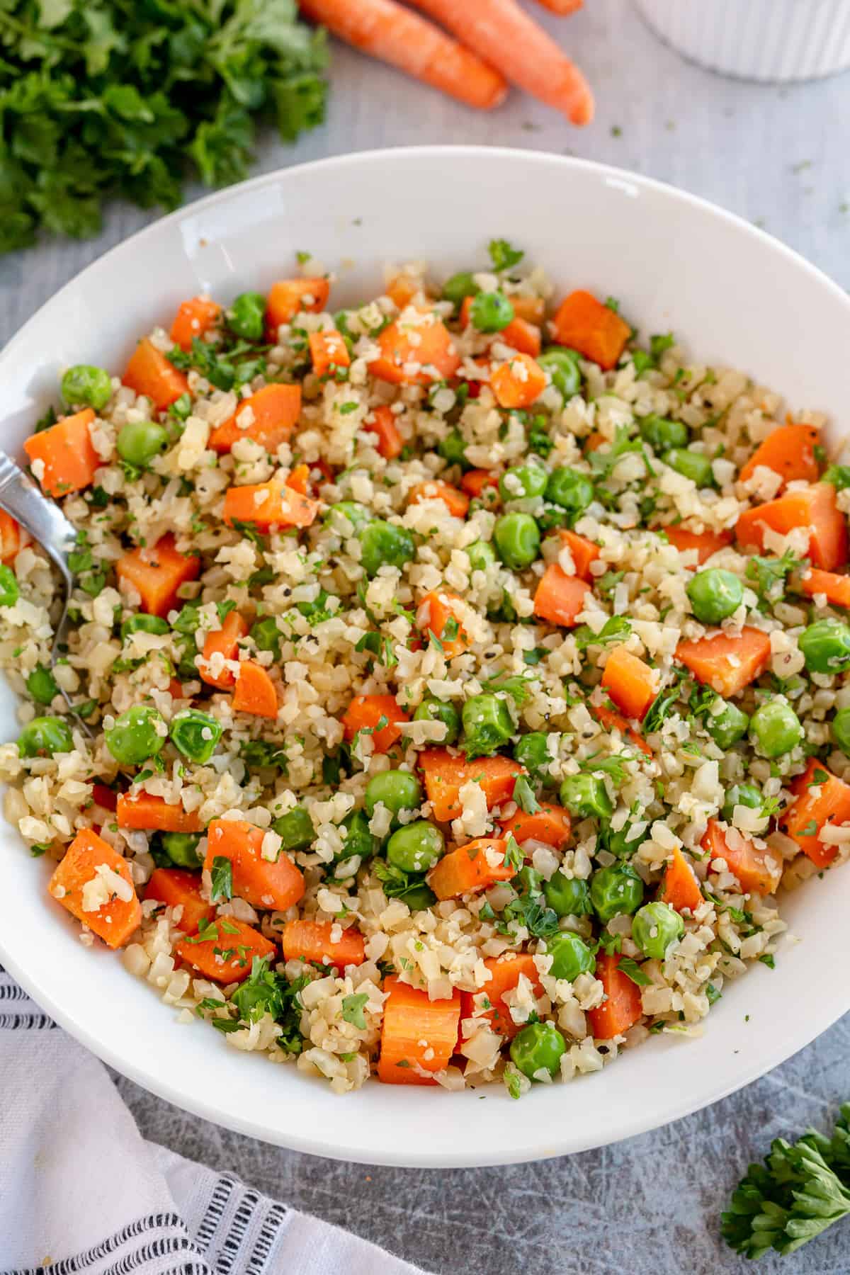 A white serving bowl full of Cauliflower Rice Pilaf next to parsley and carrots.