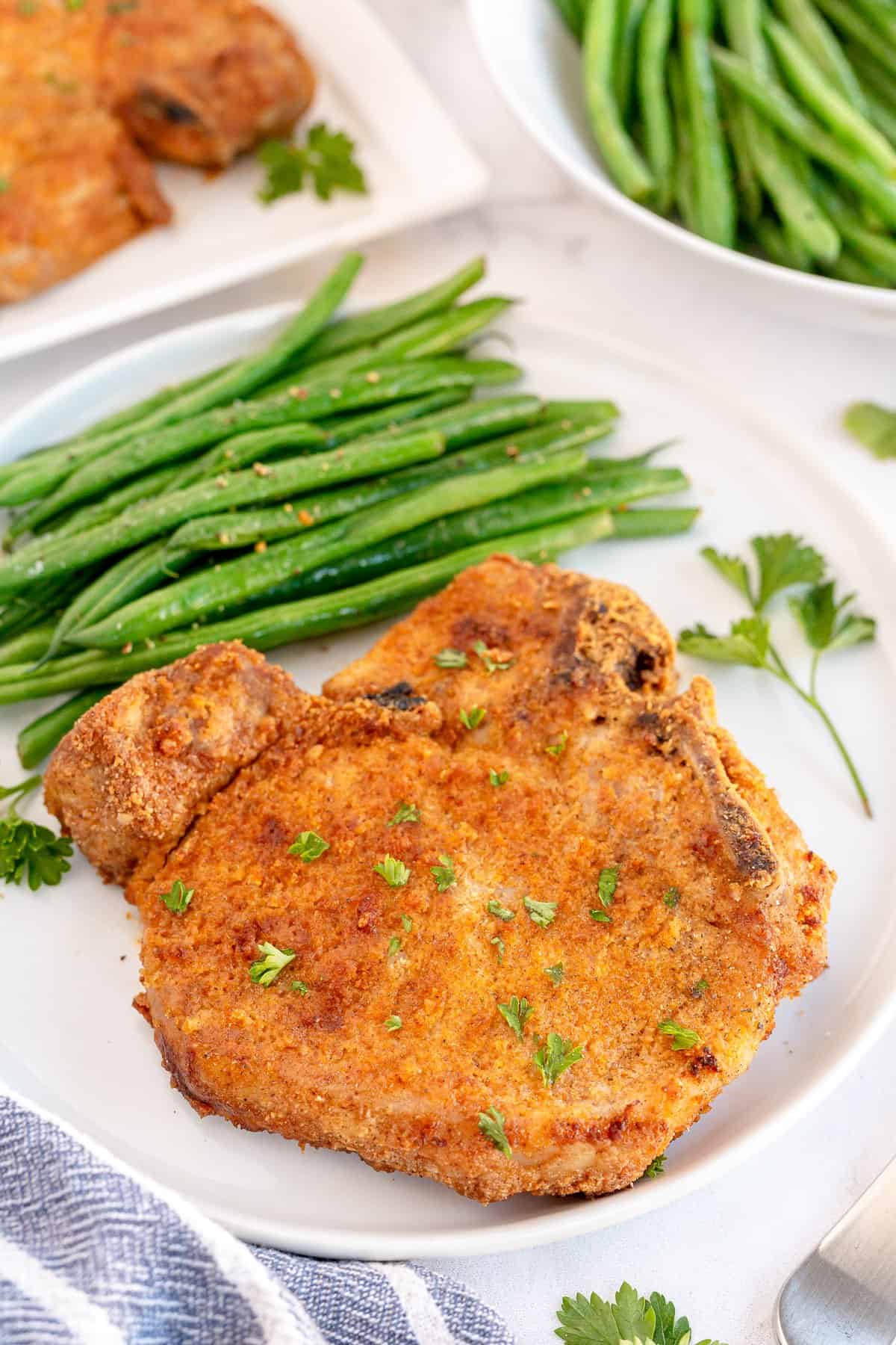 A breaded pork chop on a white plate with green beans.