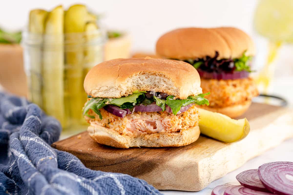Two air fried salmon sandwiches on a cutting board with a pickle.