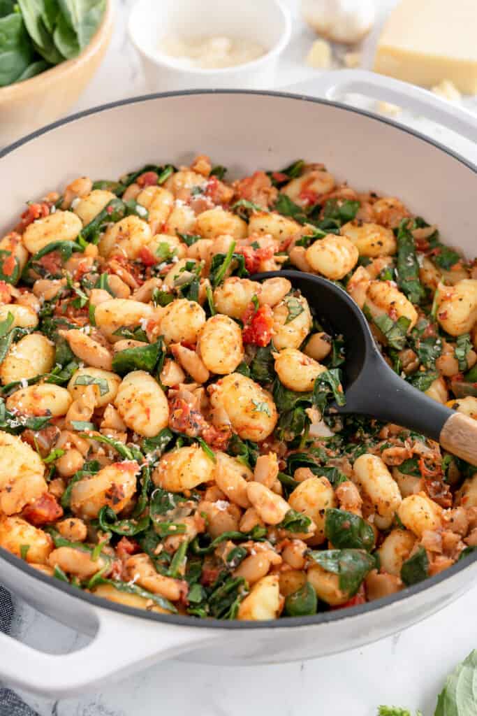 A white skillet filled with gnocchi, white beans, spinach and tomatoes.