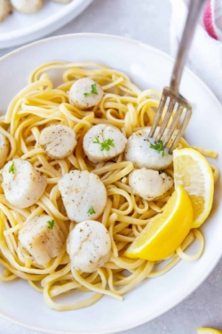 A fork piercing a scallop in a bowl with pasta and lemon.