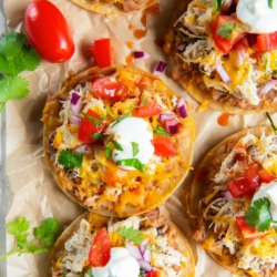 A top down shot of mini chicken tostadas on parchment paper surrounded by cilantro and grape tomatoes.