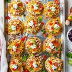 A top down shot of little tostadas with cheese, sour cream and tomatoes on a foil lined baking sheet.