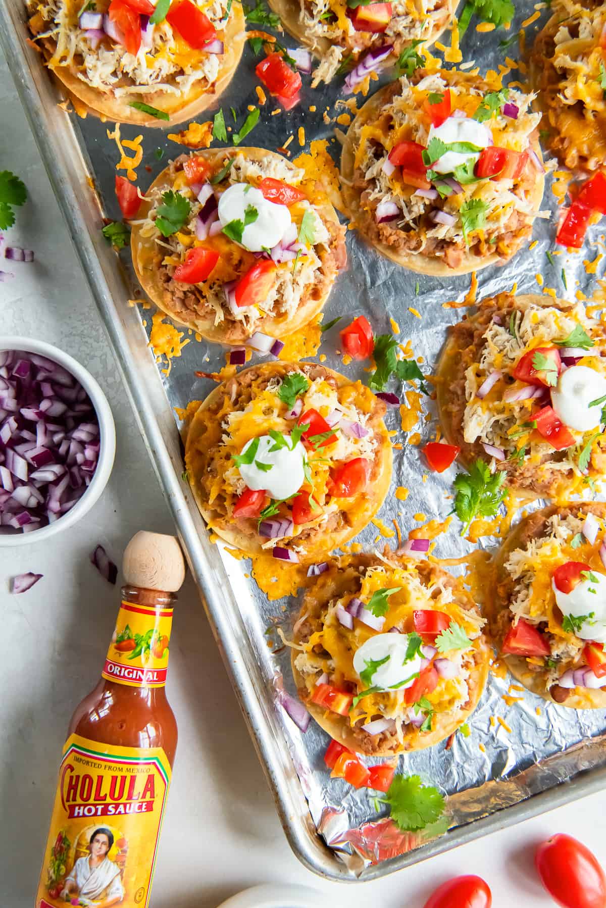 A top down shot of little tostadas with cheese, sour cream and tomatoes on a baking sheet next to a bottle of Cholula hot sauce.