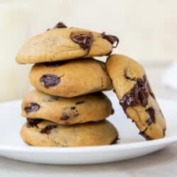 A chocolate chip cookie leaning against a stack of cookies on a plate.
