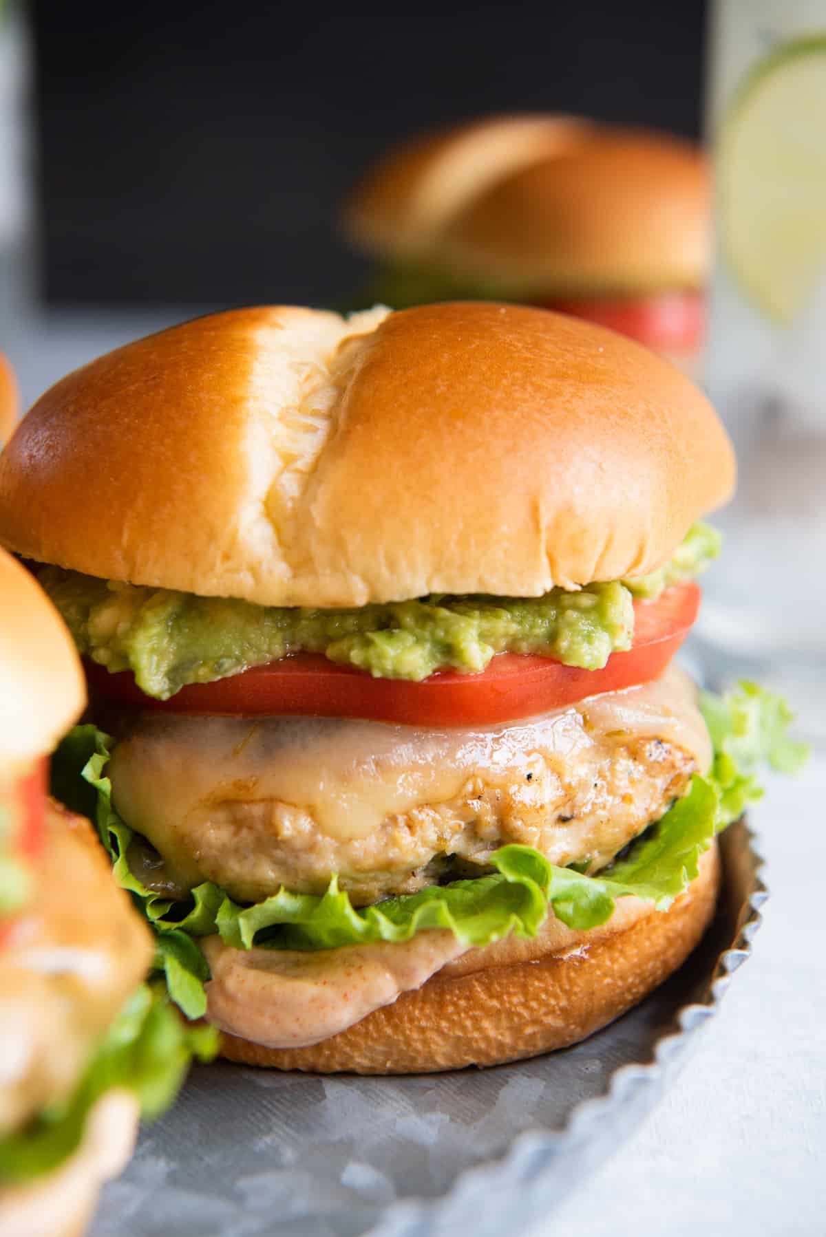 A side view of a chicken burger with sauce, lettuce, tomato, and avocado on a metal tray.
