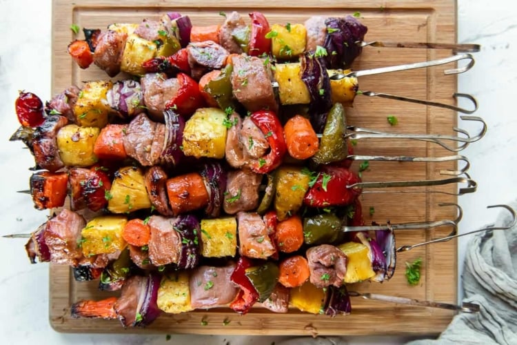 Pork Kabobs with pineapple, carrots, bell peppers, and onion piled on a wood cutting board.