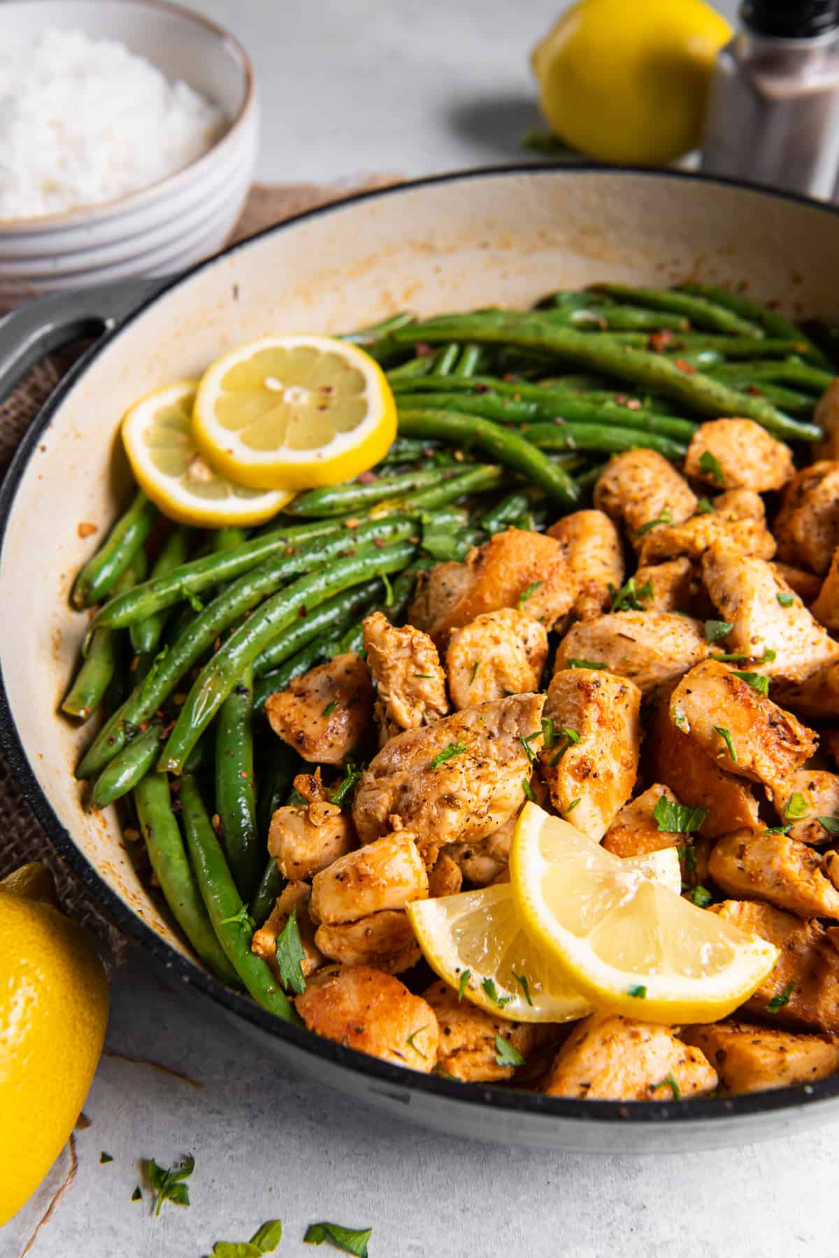 A side view of cooked chicken chunks and green beans in a skillet with lemon slices.