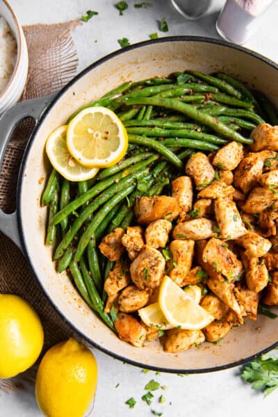 A top down shot of cooked chicken chunks and green beans in a skillet with lemon slices.