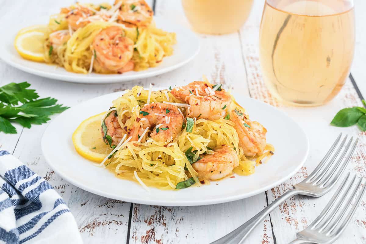 A serving of spaghetti squash shrimp scampi on a white plate with lemon slices and a fork with a glass of wine in the background.
