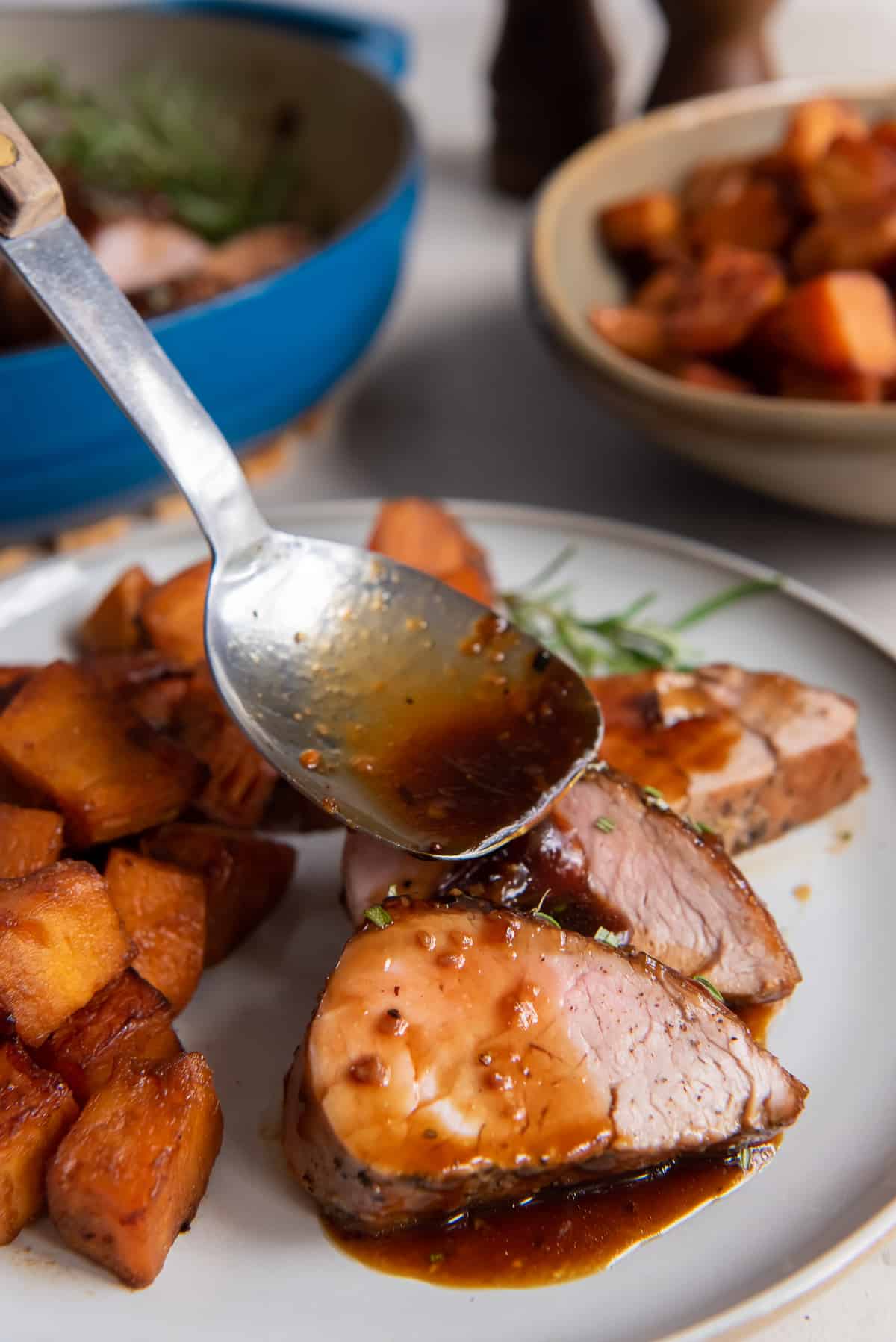 Glaze pours from a spoon over slices of pork tenderloin on a white plate.