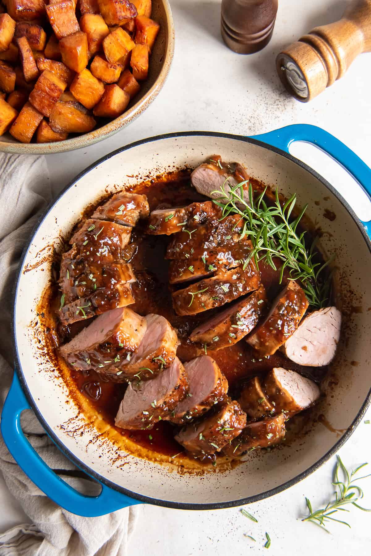A top down shot of slices of Apricot Glazed Pork Tenderloin in a pan with sprigs of fresh rosemary next to a bowl of roasted sweet potatoes and salt and pepper shakers.