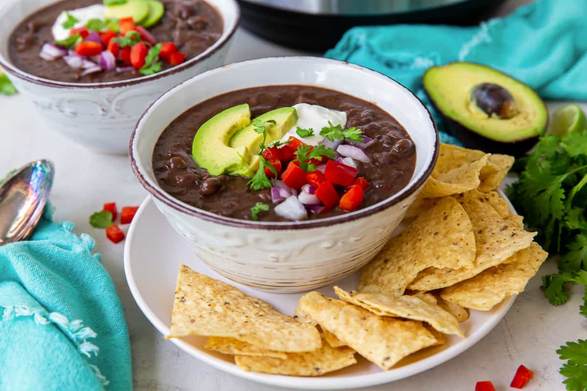 Two bowls of black bean soup topped with avocado and sour cream on plates with tortilla chips.