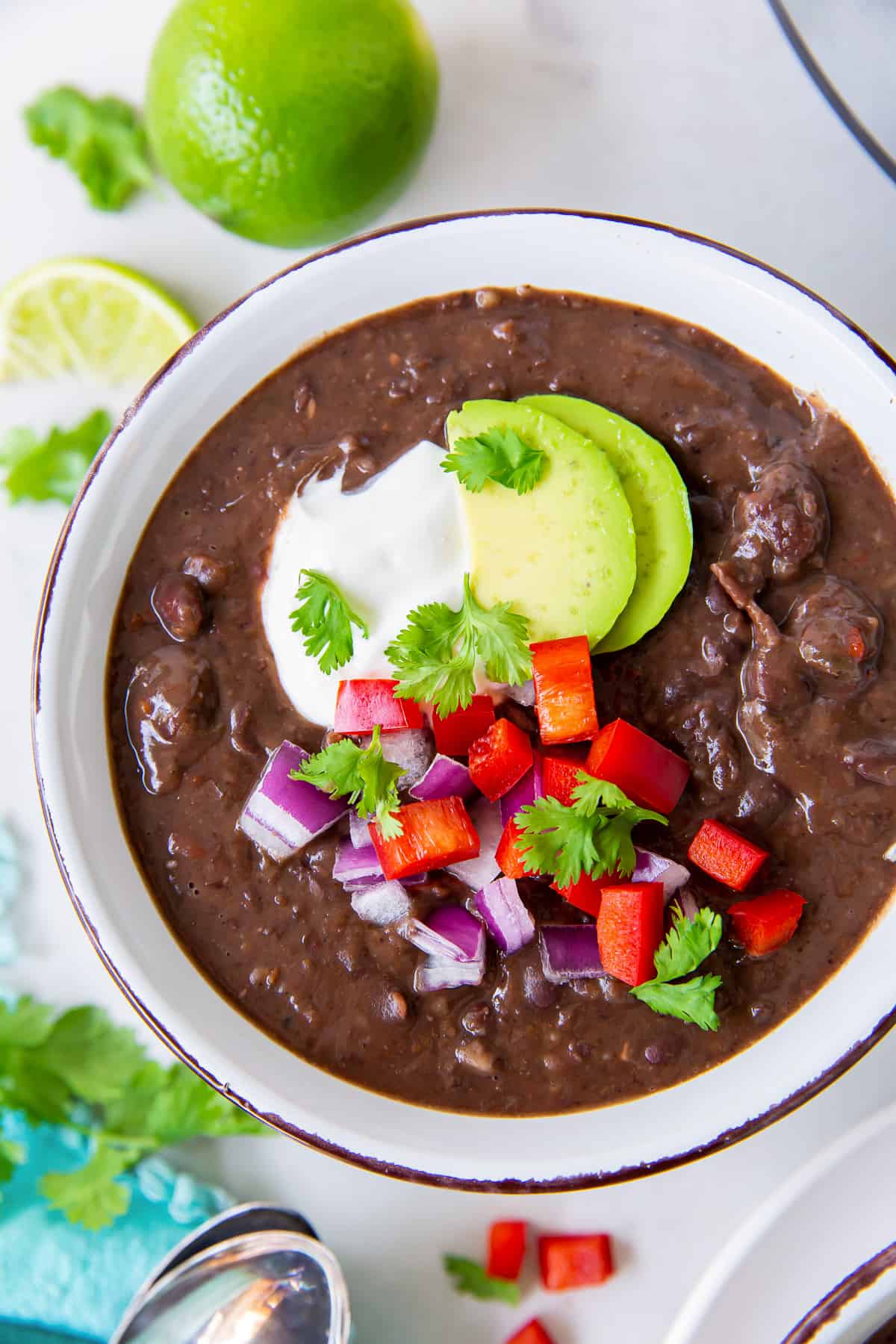 A top down shot of black bean soup topped with avocado, sour cream, onion, and red bell pepper in a white bowl.