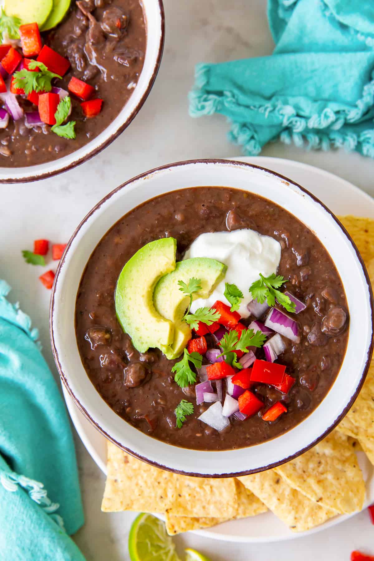 A top down shot of two bowls of black bean soup topped with avocado, sour cream, onion, and red bell pepper.