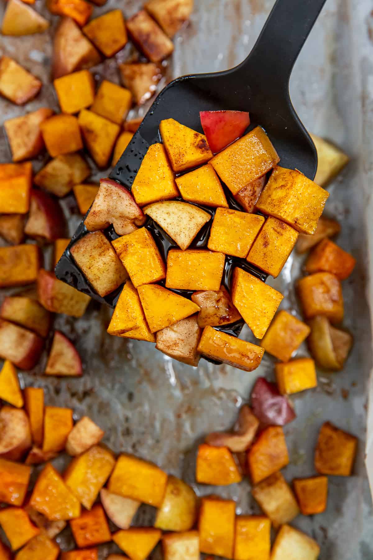 A spatula lifts roasted butternut squash and apples from a baking sheet.