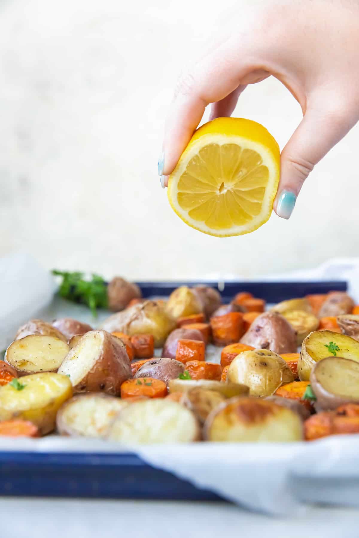 A hand squeezing juice from a lemon over a baking sheet filled with roasted carrots and potatoes.