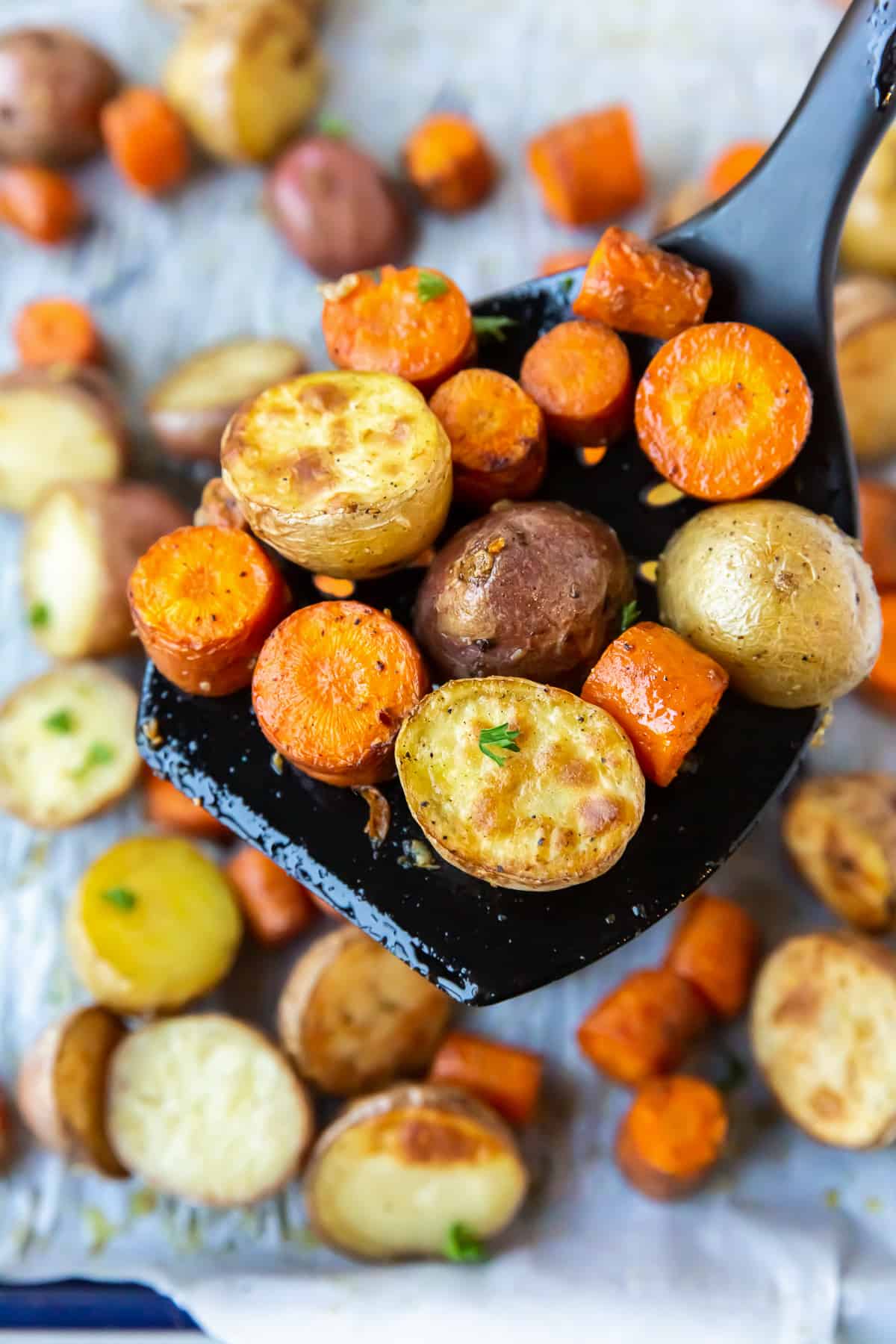 A large black spatula lifts roasted carrots and potatoes from a baking sheet.