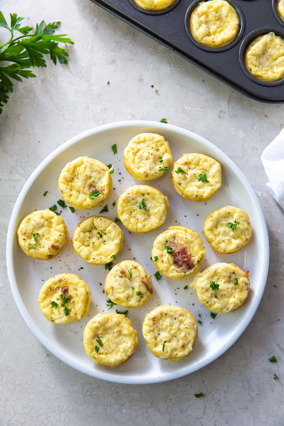 A top down shot of egg bites on a white plate next to a muffin pan and parsley.