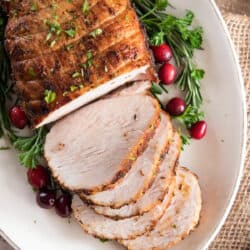 A sliced boneless turkey breast a white platter decorated with fresh herbs and cranberries.