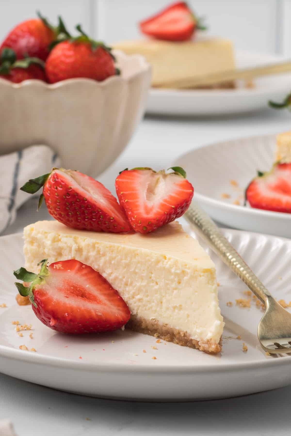 A slice of cheesecake top in front of a bowl of strawberries.ed with strawberries on a white plate with a fork.