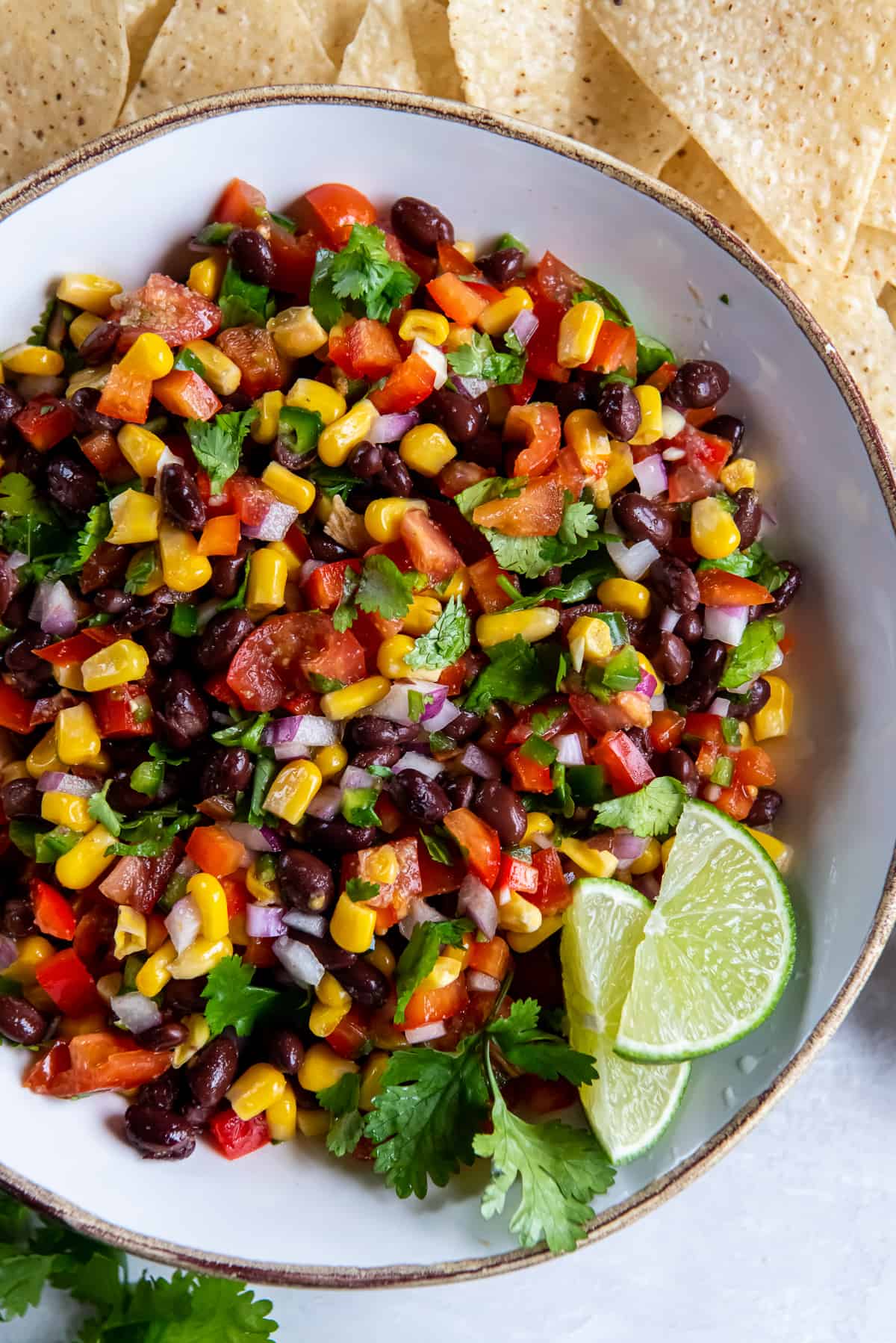 A top down shot of black bean and corn salsa in white bowl with lime wedges and cilantro.