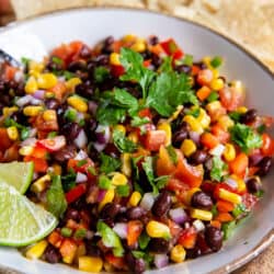 A white bowl holding black bean and corn salsa garnished with cilantro and lime wedges with tortilla chips scattered around it.