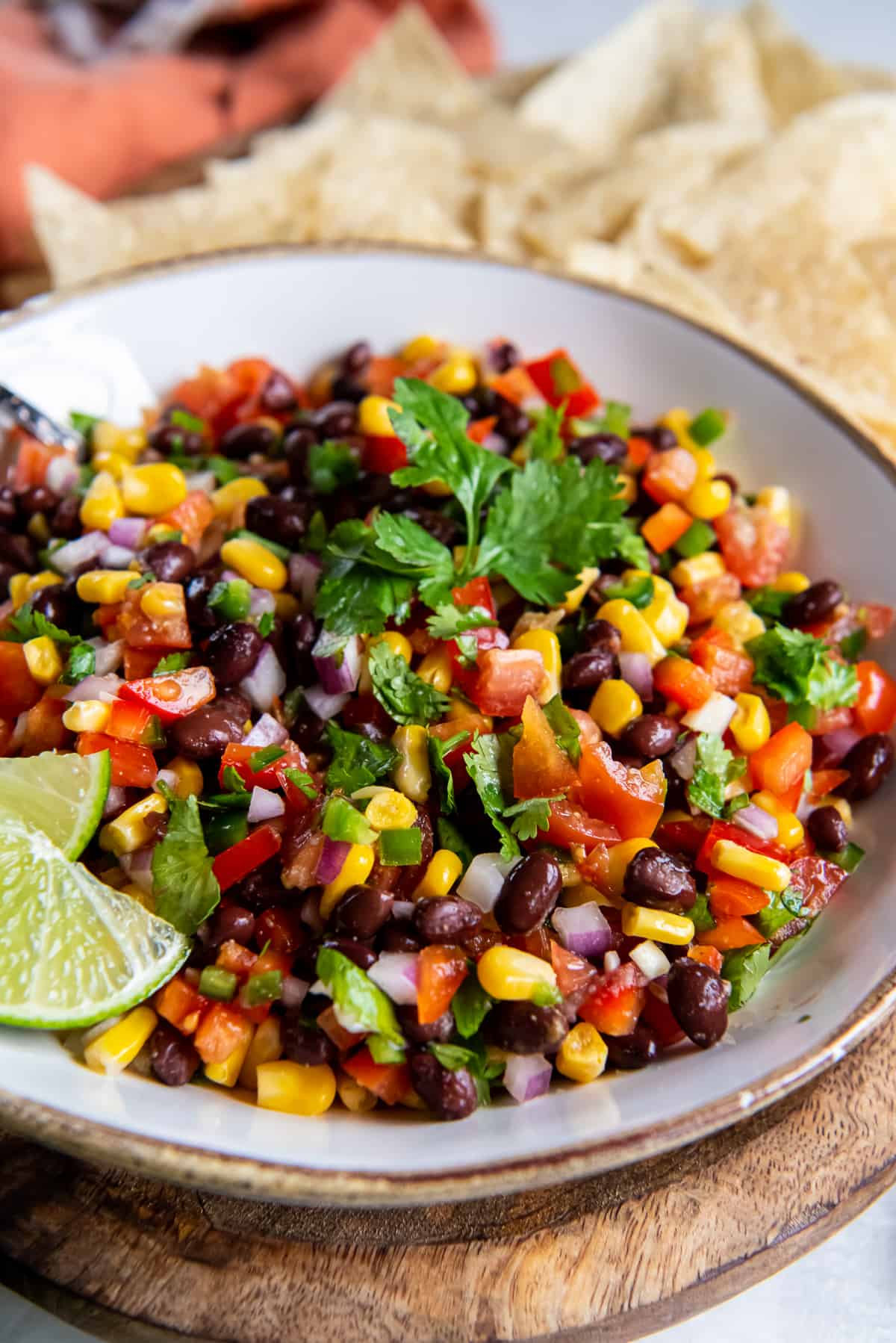 A white bowl holding black bean and corn salsa garnished with cilantro and lime wedges with tortilla chips scattered around it.