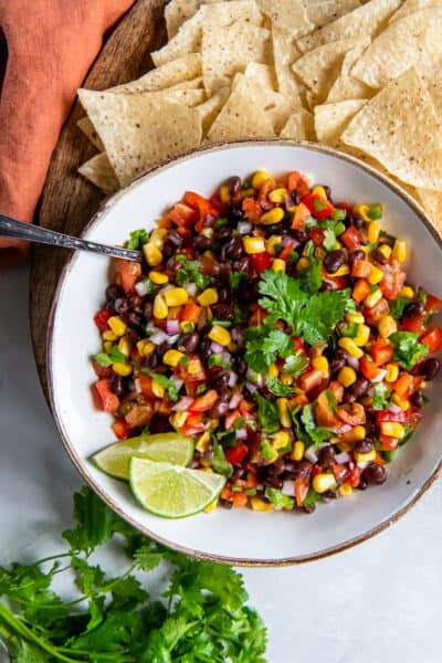 A top down shot of black bean and corn salsa in a white bowl surrounded by tortilla chips and cilantro.