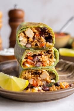 A stack of halved burritos in green tortillas revealing the filling of rice, black beans, red bell peppers, and chicken.