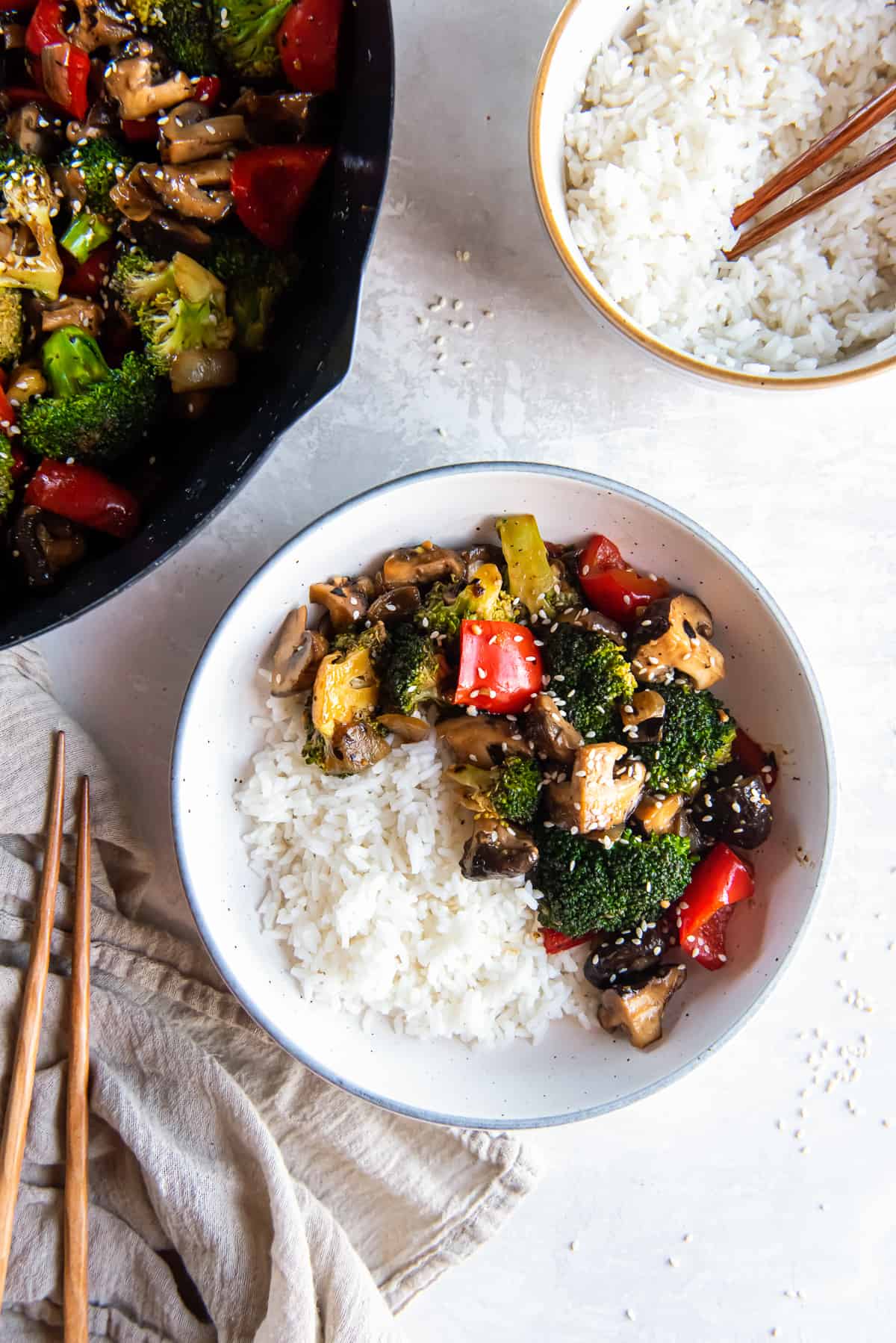 A top down shot of broccoli mushroom stir fry with white rice in a bowl next to a wok and a bowl of rice.