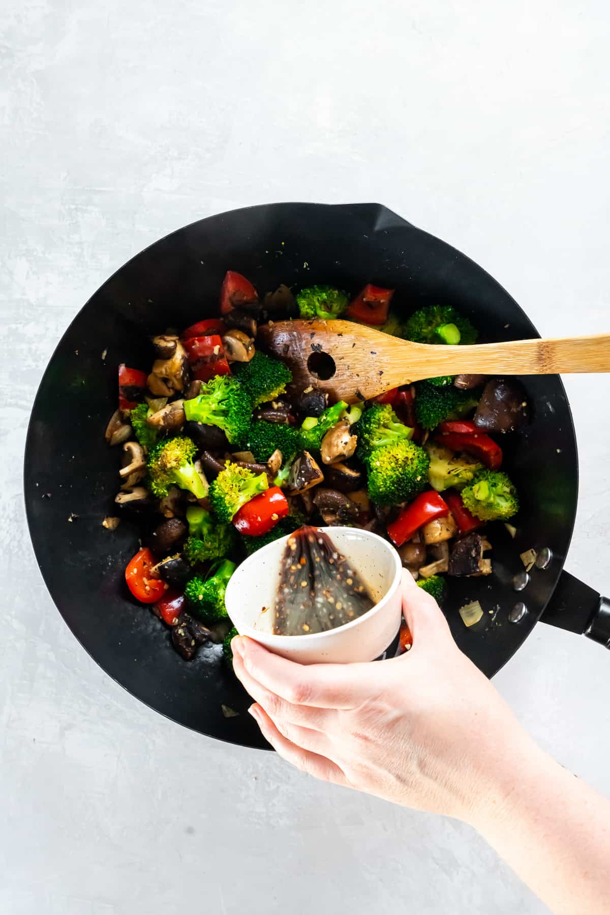 A hand pouring stir fry sauce from a small bowl into vegetables in a wok.