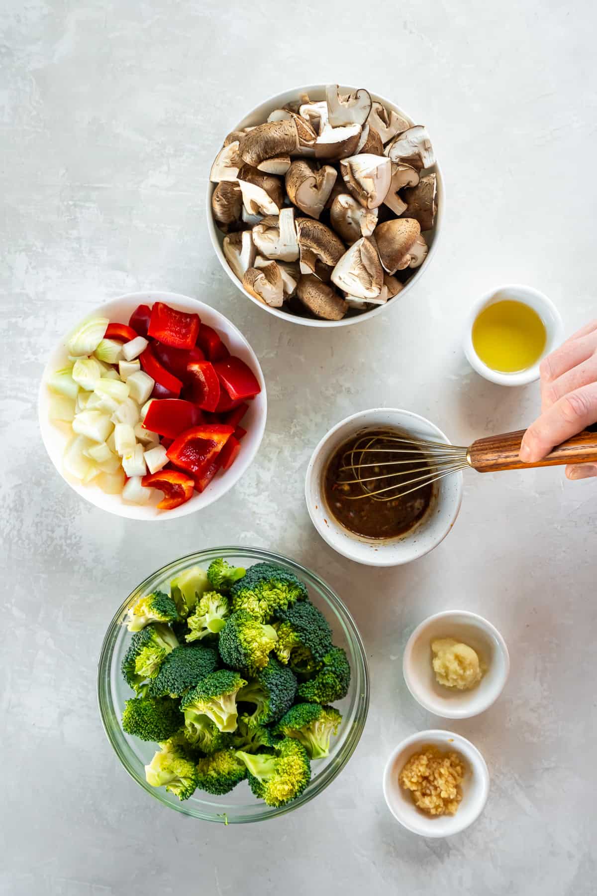 A hand whisking stir fry sauce in a small bowl surrounded by broccoli, mushrooms and other ingredients in bowls.